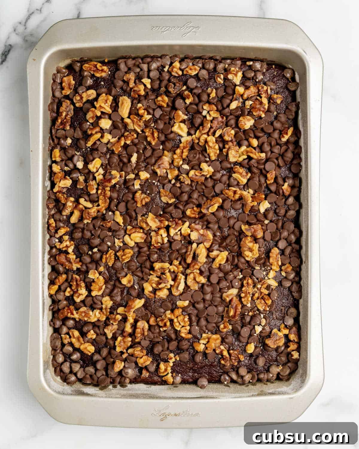 Decadent Chocolate and Date Cake Recipe for Moist Loaves 10 Metal rectangular cake pan with walnut and chocolate chip topped cake. White marble surface.