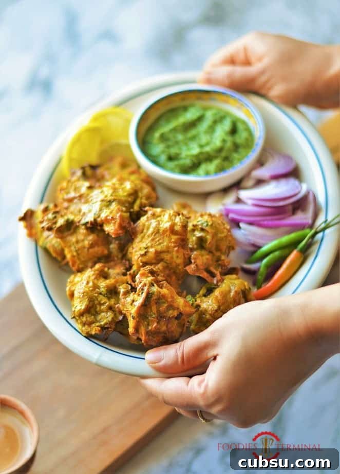 Chicken Pakora served in an oval white plate with chilies, onion slices, cilantro chuntey & lemon slices.