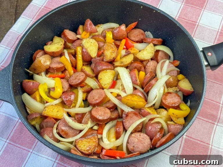 Deep skillet full of an easy Kielbasa recipe with delicious wedges of potatoes, slices of onions and pieces of bell peppers with seasoning on a red and white plaid napkin.