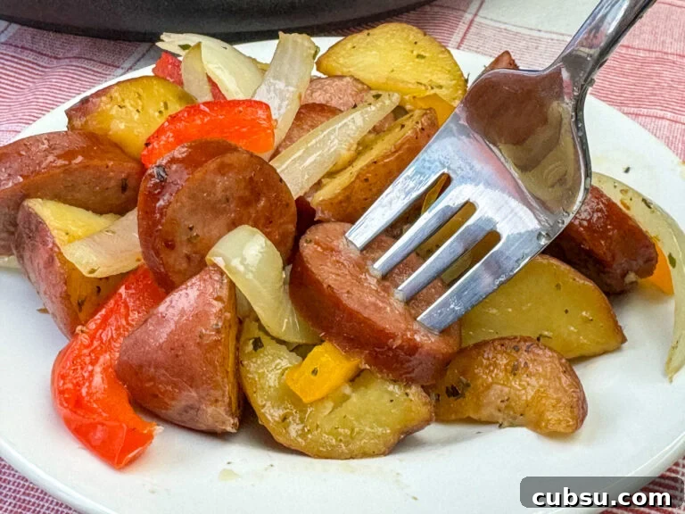 Close up of a fork in a slice Kielbasa sausage, onion and potato wedge on a plate full of the recipe.