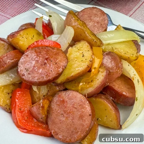 Kielbasa and potatoes with flavorful sausage and generous seasoning paired with slices of onions and peppers for additional flavor accents on a white plate with a fork in the background.