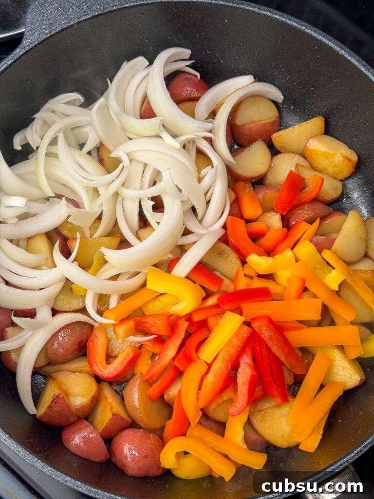 Raw slices of onions and red, yellow and orange bell peppers added to a non-stick skillet with partially cooked potatoes.