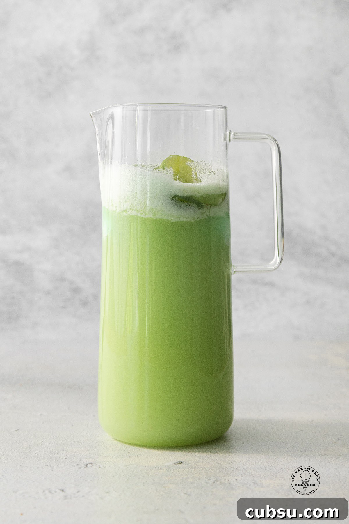 a tall glass pitcher filled with green lime sherbet punch with foam on top.