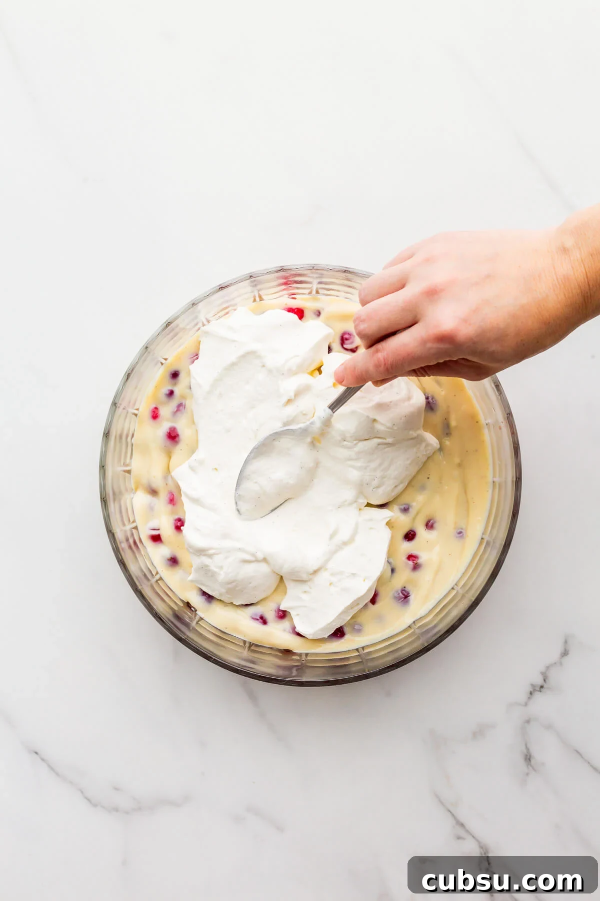 Spreading whipped cream over a layer of pastry cream to make trifle in a glass bowl.