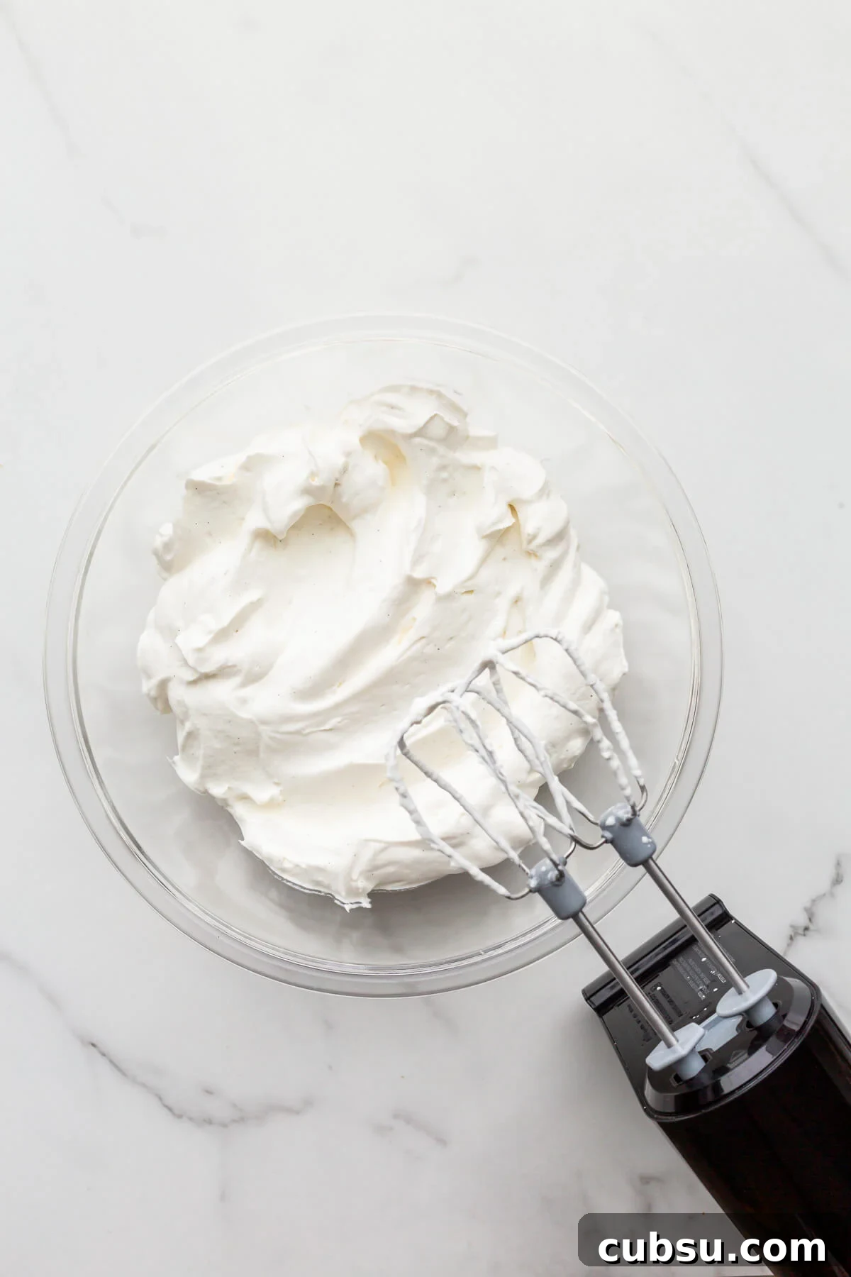 A glass bowl of vanilla whipped cream made with an electric hand mixer.