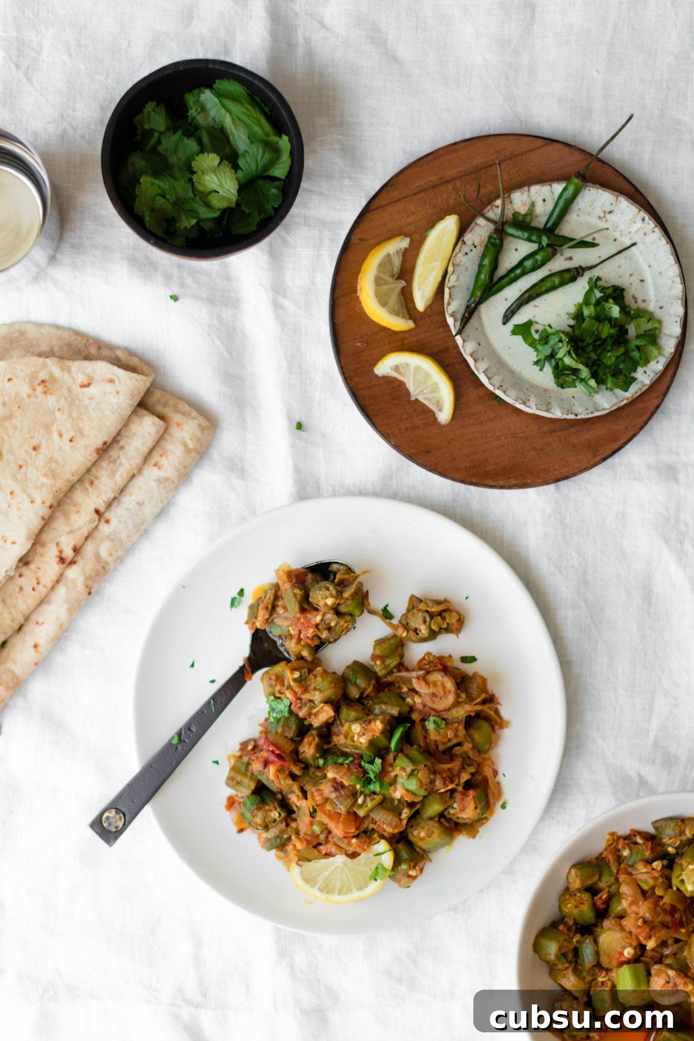Okra curry on a white plate with black spoon and served with roti and lemons on the side