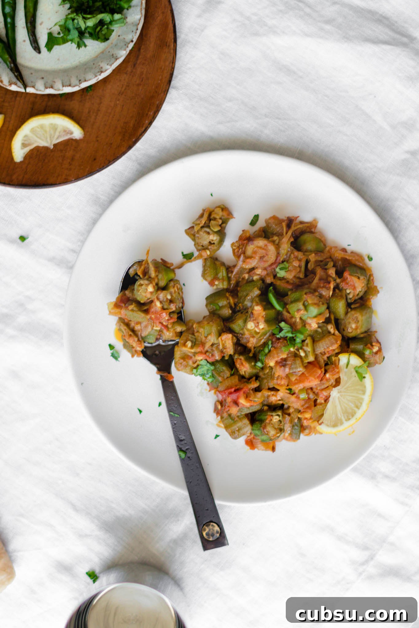 Close up of cooked okra curry on a white plate with a lemon wedge and black spoon