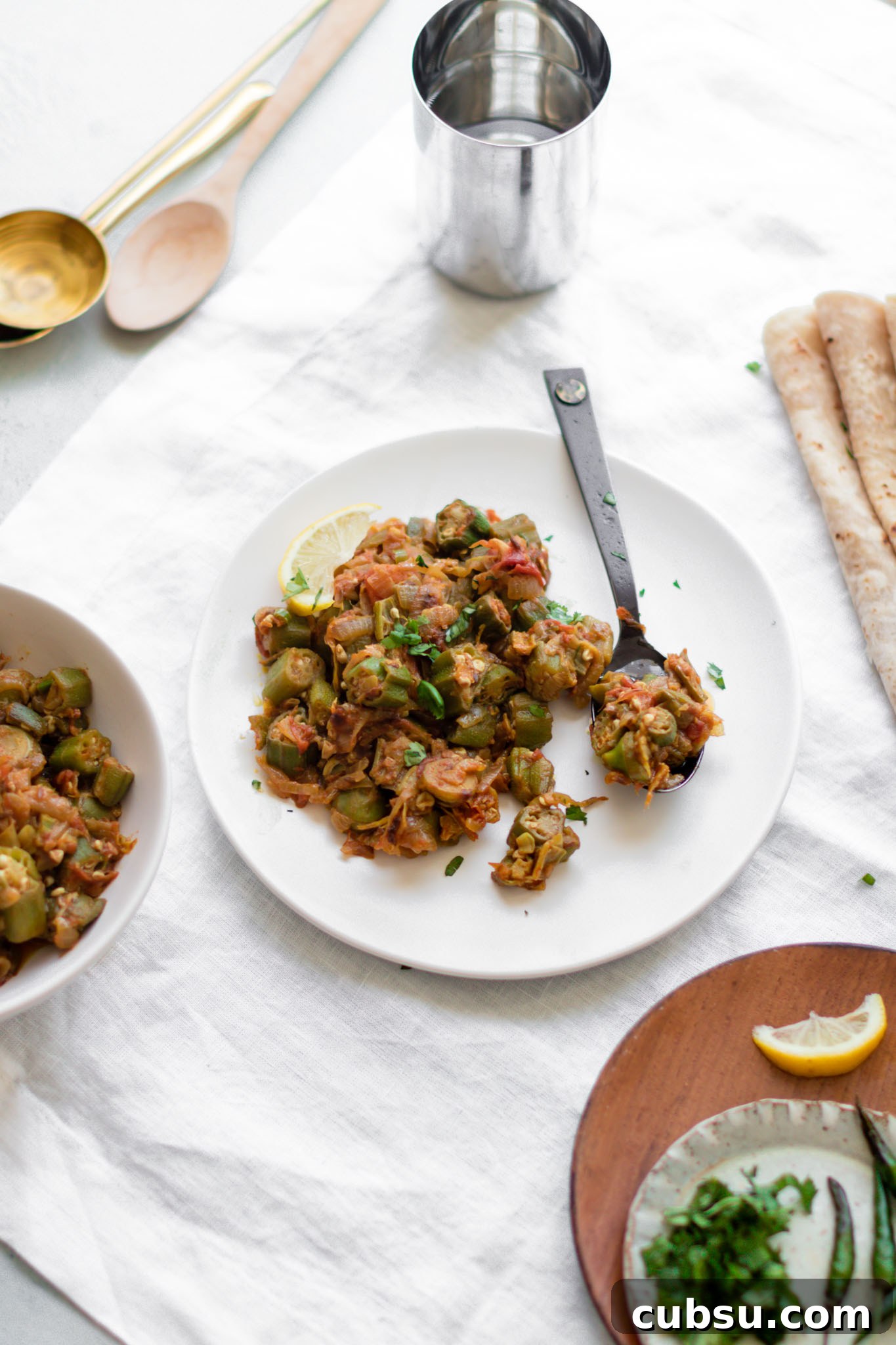 Bhindi Masala (Okra Curry) on a white plate with spoon and a cup, roti, and garnishing on the side
