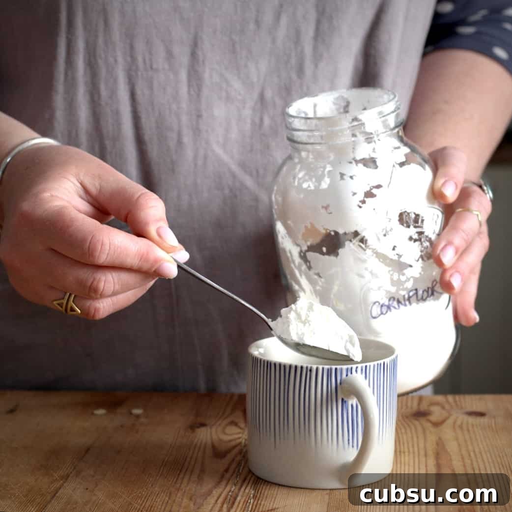 Slow Cooker Pork in Cider and Mustard Sauce 13 Woman in grey spooning a heaped teaspoon of cornflour from a glass jar into a stripy blue and white mug