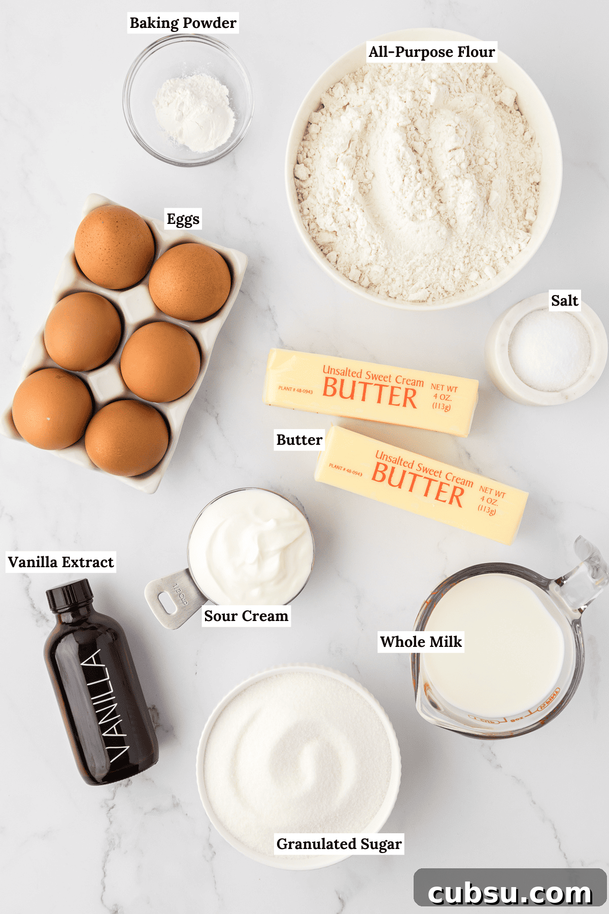 overhead view of the ingredients for bundt cake including a small glass bowl of baking powder, a white bowl of all-purpose flour, a carton of 6 eggs, a small white bowl of salt, two sticks of butter, a bottle of vanilla extract, a small measuring scoop of sour cream, a glass measuring cup of whole milk, and a white bowl of granulated sugar