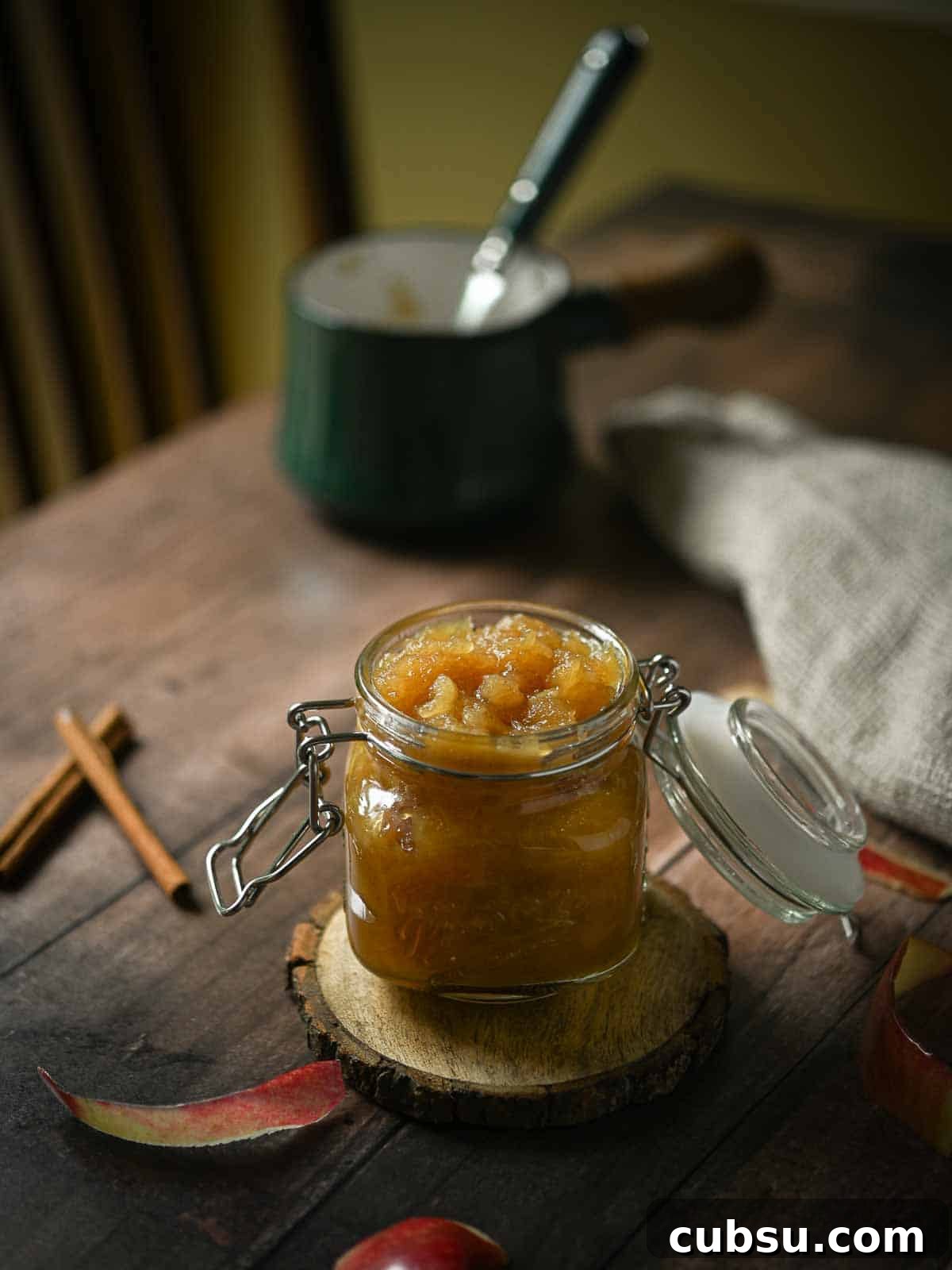 A moody shot of Honeycrisp applesauce in a clear jar on a wooden table with a pot in the background.