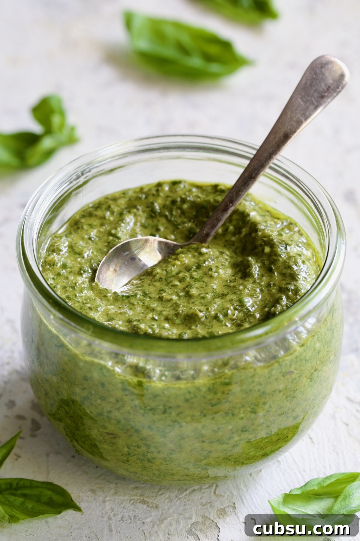 Jar of Pesto Genovese with spoon with fresh basil leaves scattered about.
