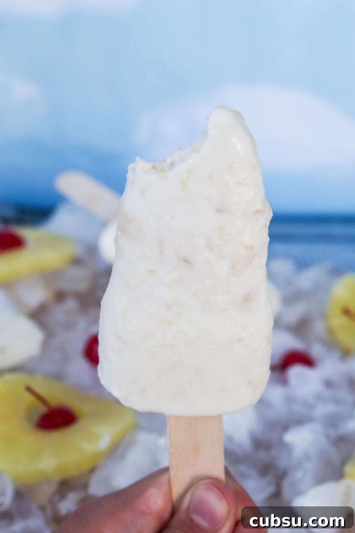 pina colada popsicle, with a bite taken out of it, being held up by hand and ice, pineapple rings, cherries and more popsicles in the background