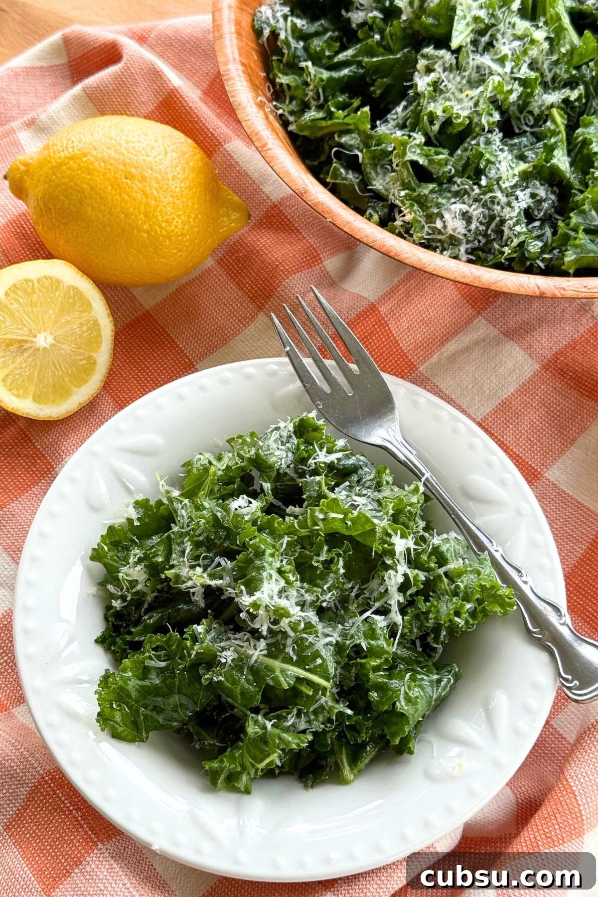 Massaged kale salad topped with finely grated Parmesan cheese and lemon dressing in a white bowl on a orange checkered background with a lemon and serving bowl with more behind it.
