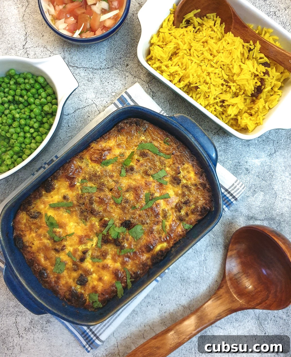 A blue serving dish of bobotie next to a bowl of yellow rice and a bowl of peas.