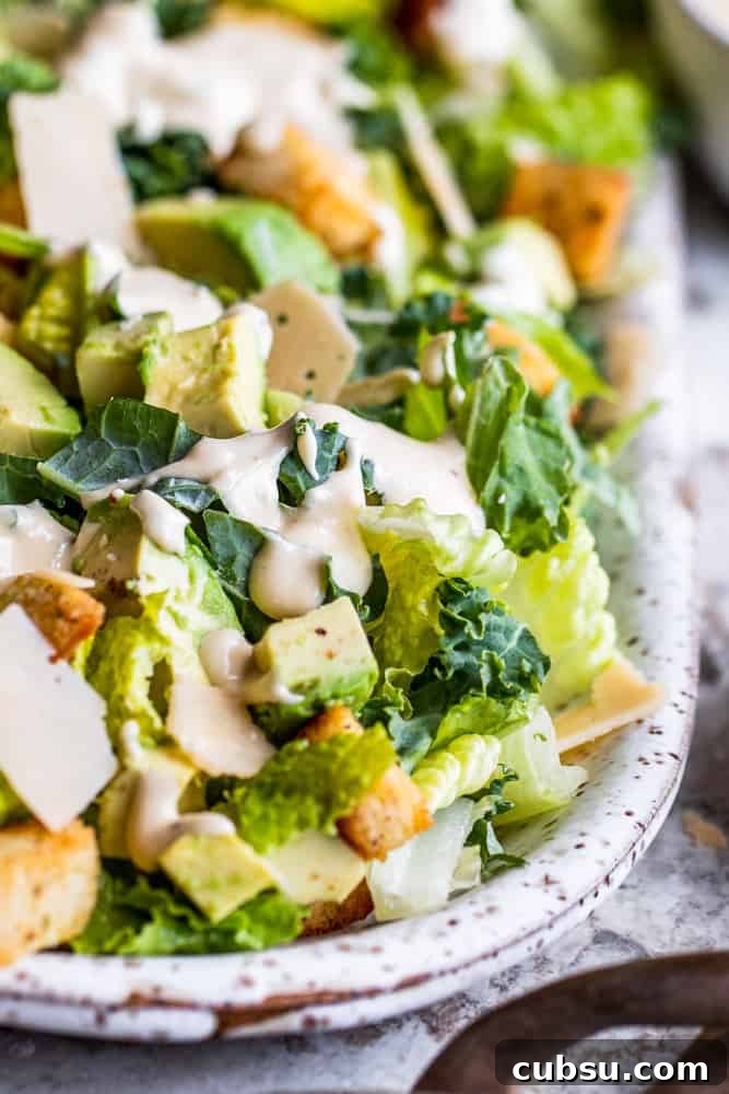 Kale Caesar Salad with avocados, homemade croutons, Caesar dressing and Parmesan cheese. Caesar salad on a white platter on a white counter.