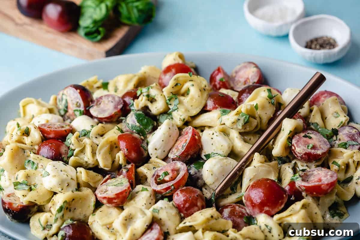tortellini caprese salad on platter next to cutting board with tomatoes and basil, and small bowls of salt and pepper