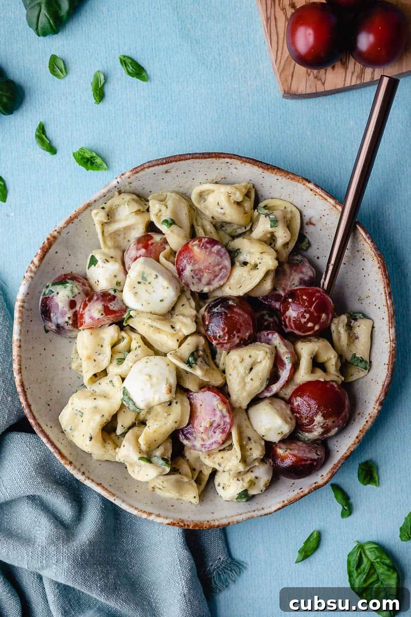 small bowl of tortellini caprese salad next to napkin, basil leaves, and cutting board with tomatoes