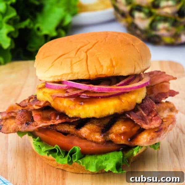 Square format image of a grilled Hawaiian chicken sandwich piled high with lettuce, tomato, bacon, pineapple, and red onion on a soft bun.