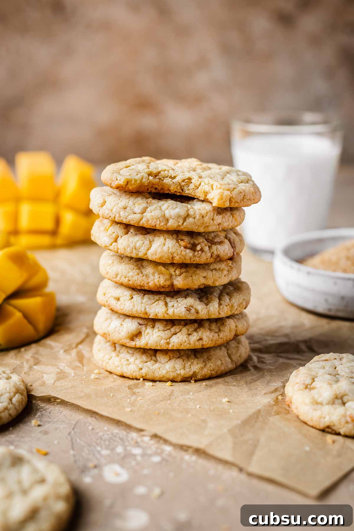 A stack of seven mango cookies on brown parchment paper with a glass of milk and sliced mangos in the background.