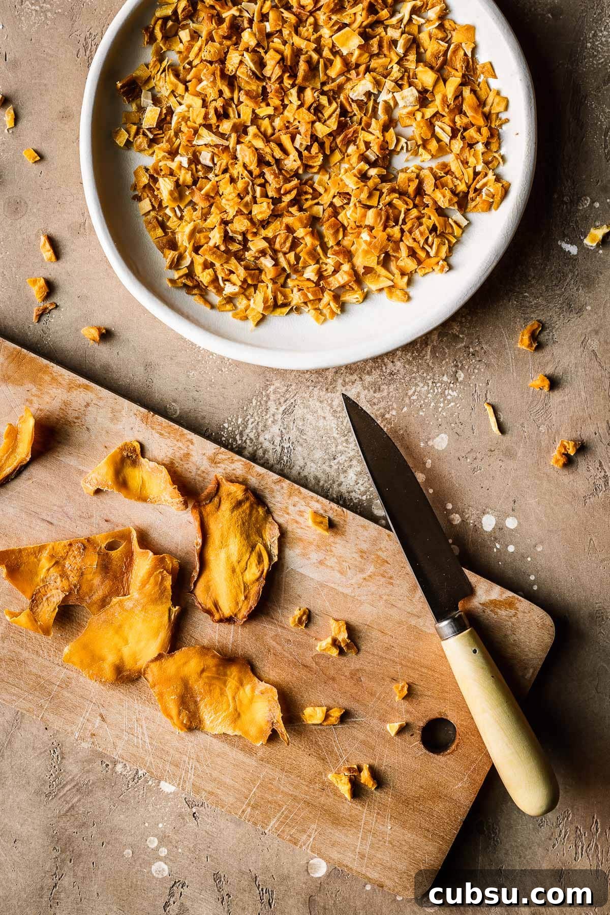 A plate of diced dried mangos alongside a wooden cutting board, paring knife and dried mango cheeks.