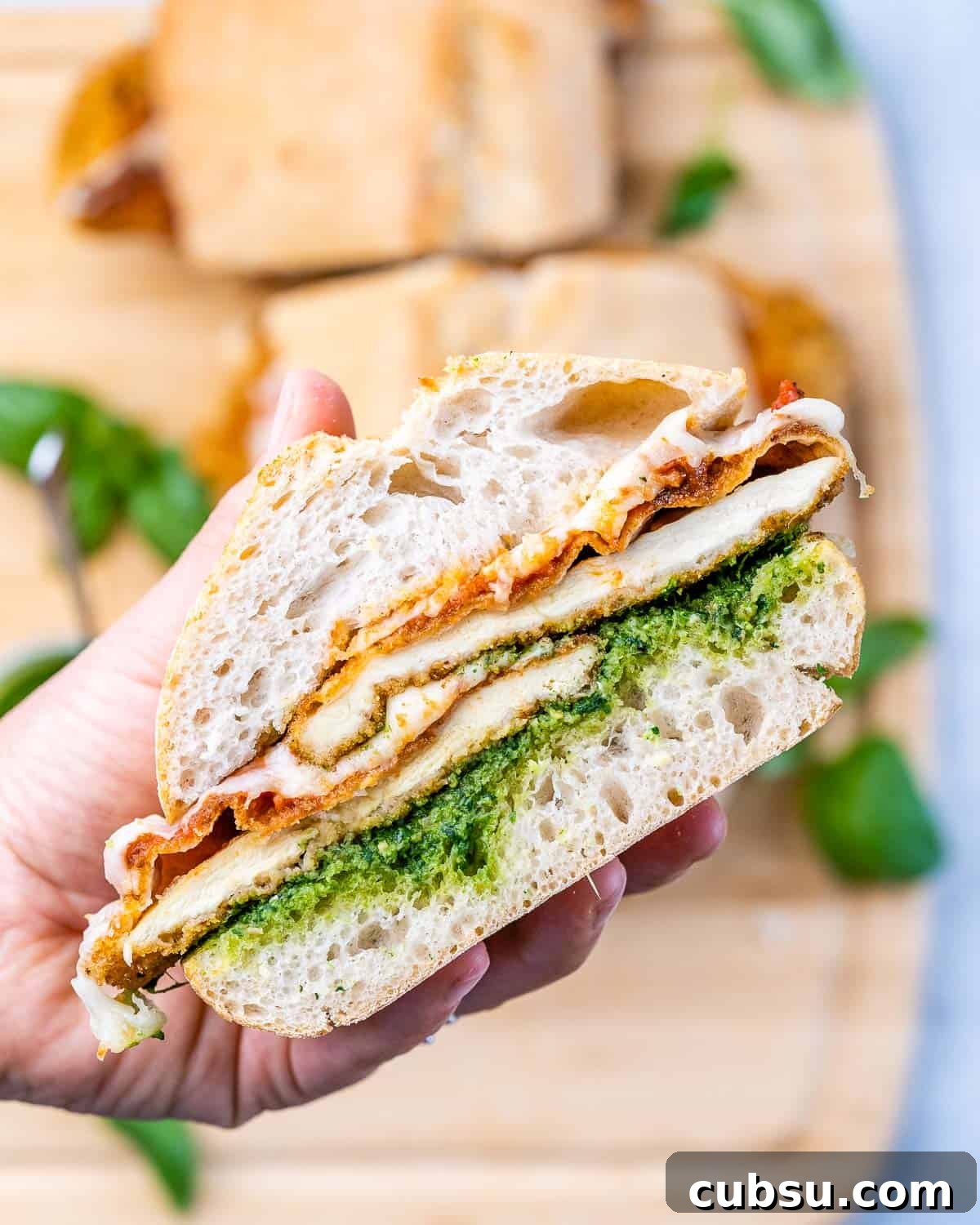 Crispy Chicken Parmesan Sandwich Recipe That Beats Takeout 7 Hand holding a Chicken Parmesan Sandwich with crispy breaded chicken, melted mozzarella, marinara, and bright green pesto on ciabatta.