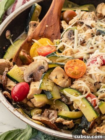 Cooked chicken, zucchini, tomatoes, and mushrooms in metal pan with wooden spoon.