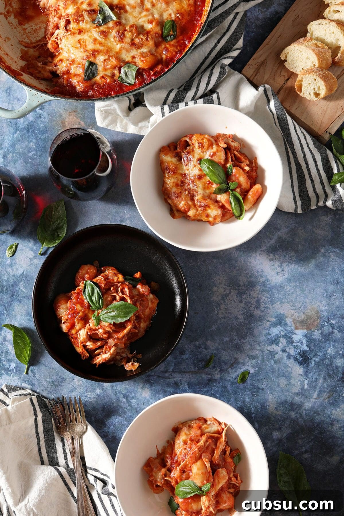 Three bowls of a gnocchi bake with chicken on a blue table with forks, wine, bread and the baking dish