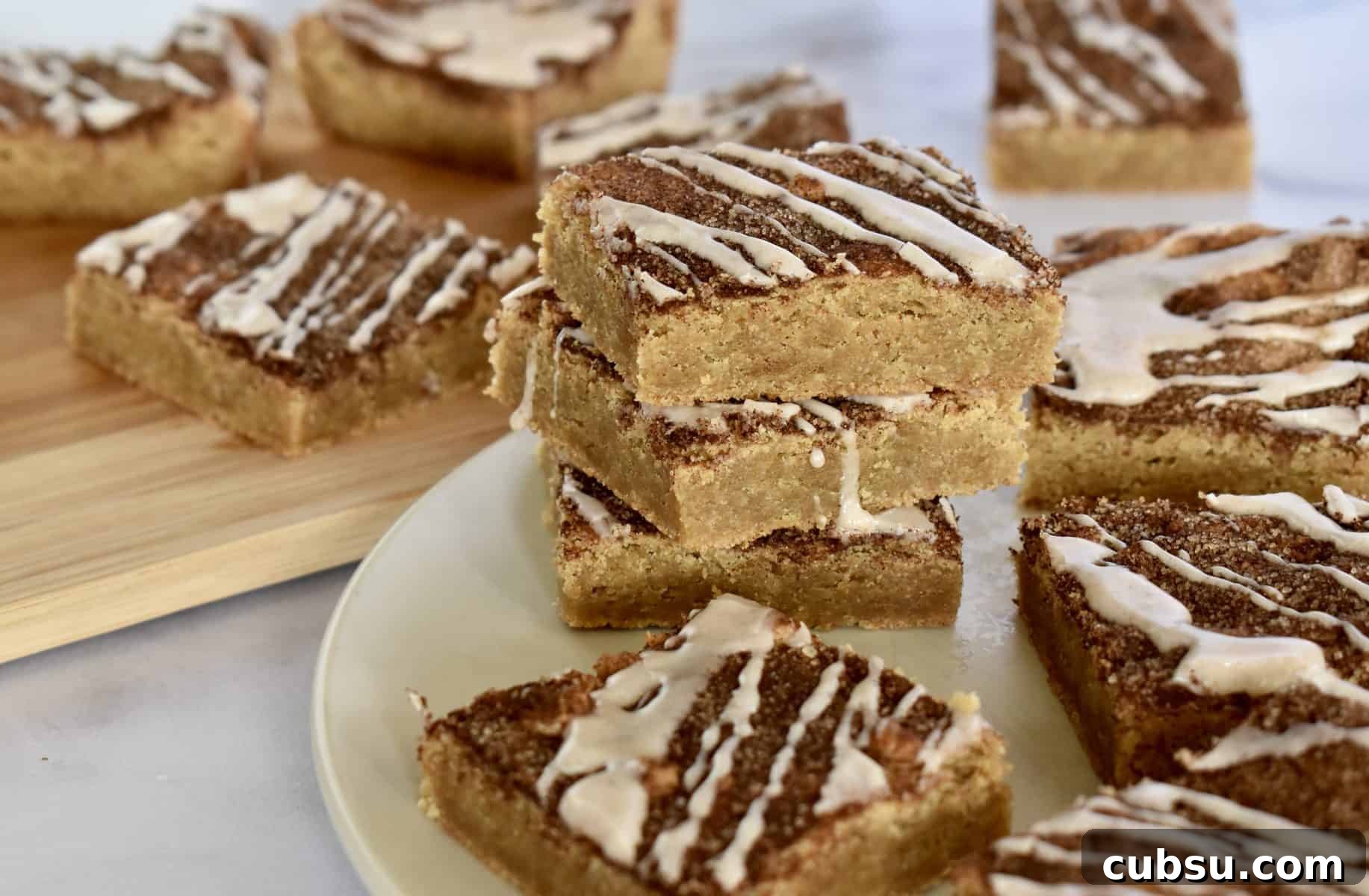 Chewy Cinnamon Sugar Blondies Recipe for Perfect Texture 12 Cinnamon sugar blondies stacked high on a white plate with glaze drizzled over top.