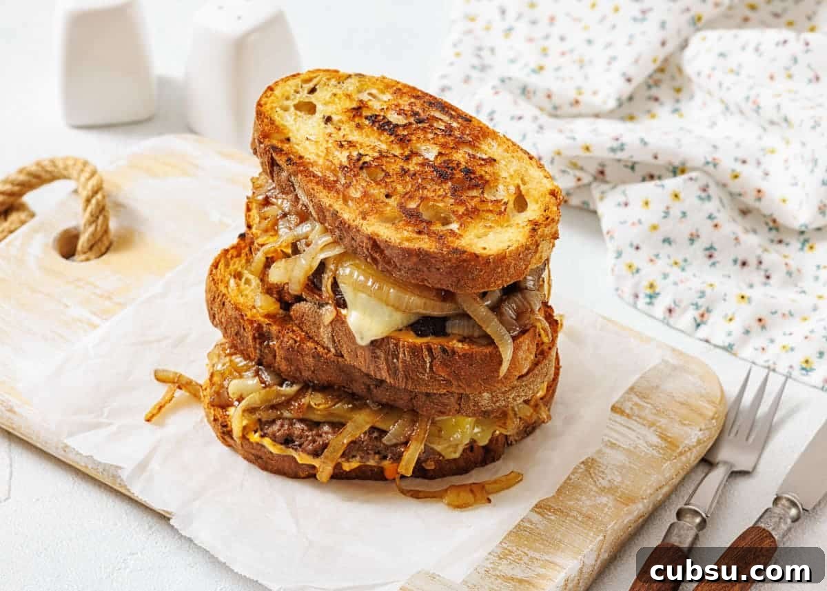 31 Classic Comfort Recipes That Keep Families Coming Back 11 A stack of two patty melt sandwiches with caramelized onions and melted cheese on toasted bread, placed on parchment paper atop a wooden board.