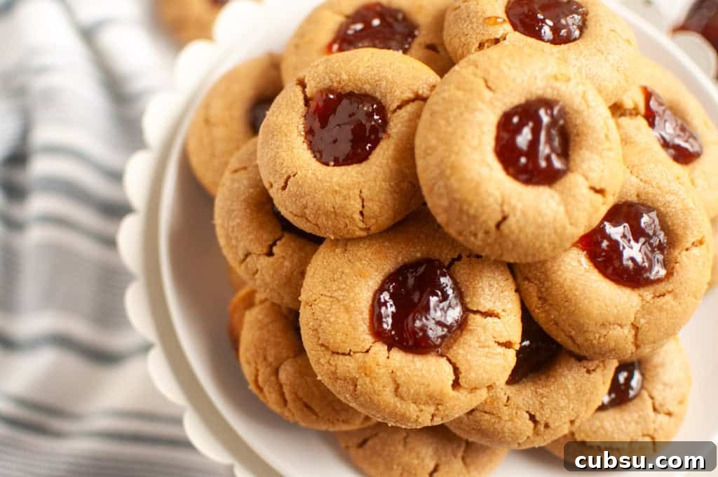 31 Classic Comfort Recipes That Keep Families Coming Back 16 A plate stacked with Peanut butter jelly thumbprint cookies filled with red jam, placed on a white stand next to a striped cloth.