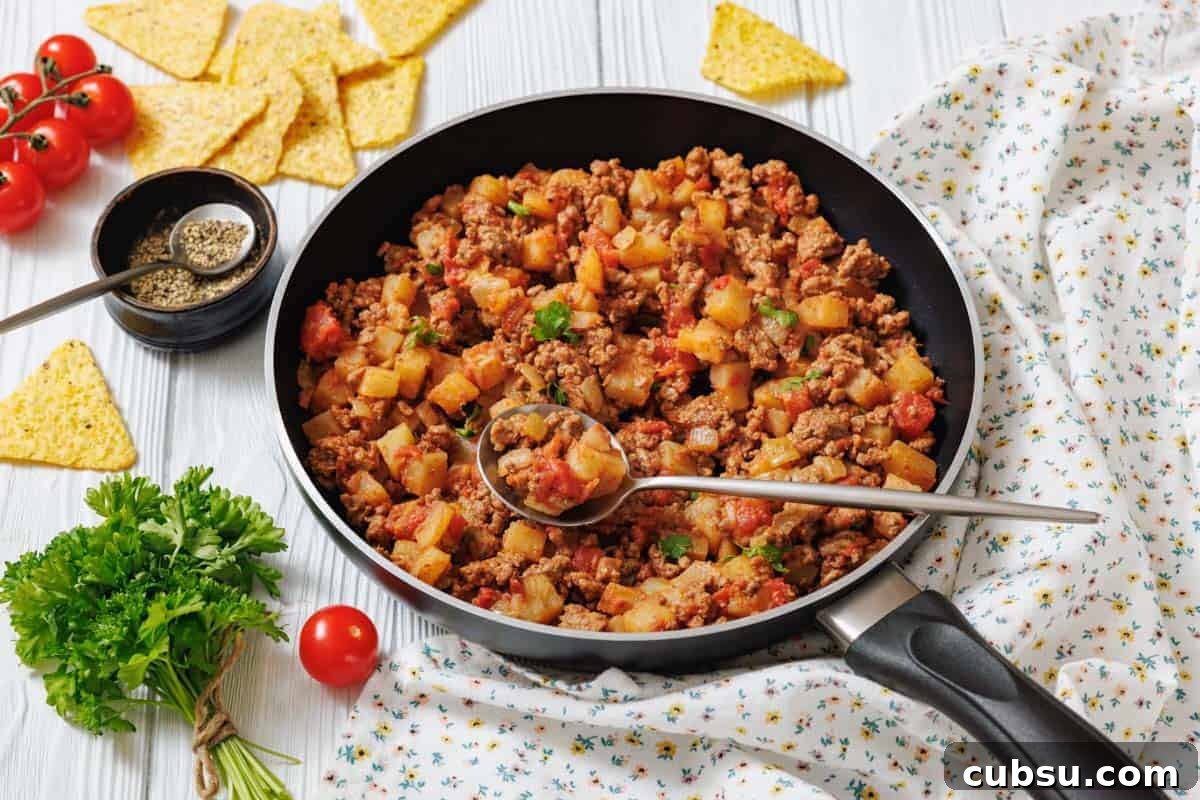 31 Classic Comfort Recipes That Keep Families Coming Back 17 A frying pan filled with Poor Man's meal sits on a white surface with a spoon, surrounded by fresh parsley, tomatoes, tortilla chips, and seasoning.