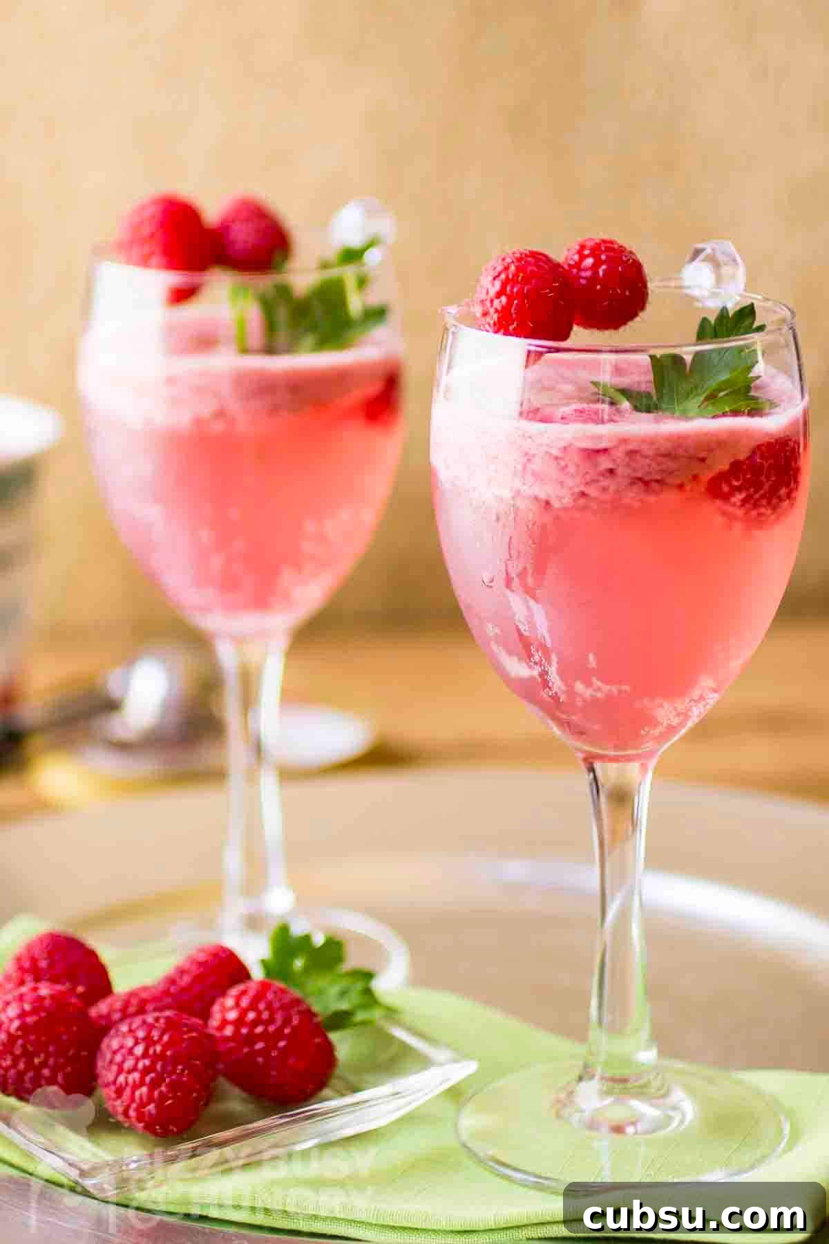 3-Ingredient Raspberry Spritz Cocktail Recipe 2 Side shot of two cocktail glasses with raspberry spritz garnished with fresh mint and raspberries with more raspberries and a green cloth underneath.