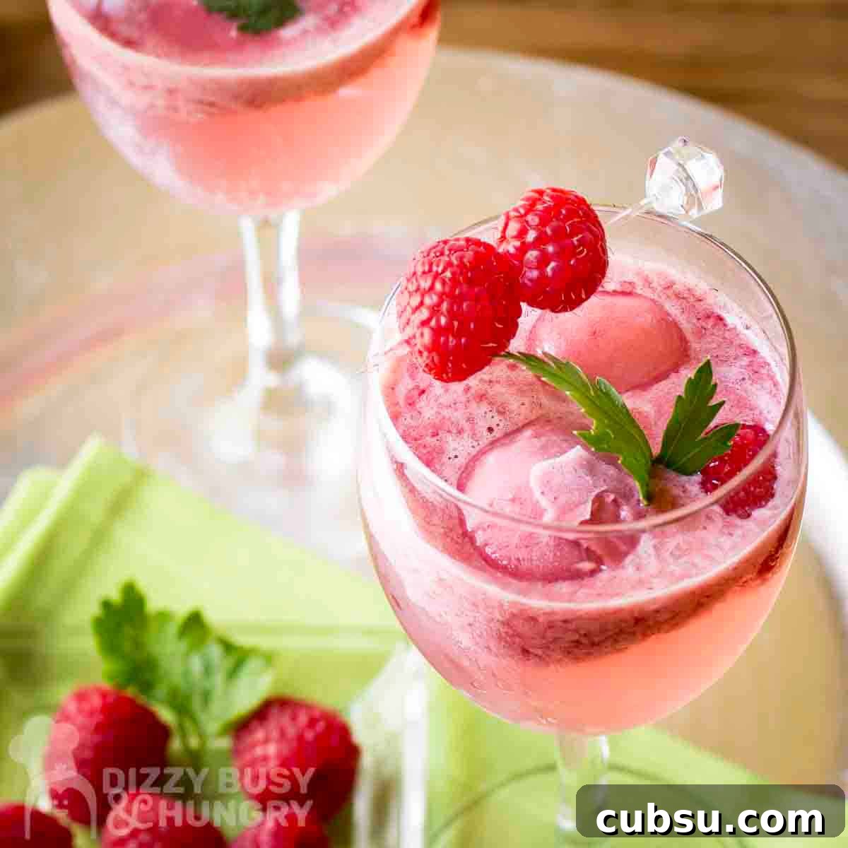 3-Ingredient Raspberry Spritz Cocktail Recipe 7 Close up shot of two cocktail glasses with raspberry spritz garnished with fresh mint and raspberries with more raspberries and a green cloth underneath.
