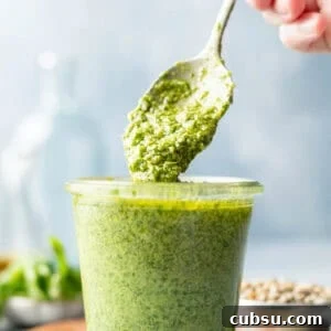 Pesto sauce without pine nuts drips from spoon into small jar.