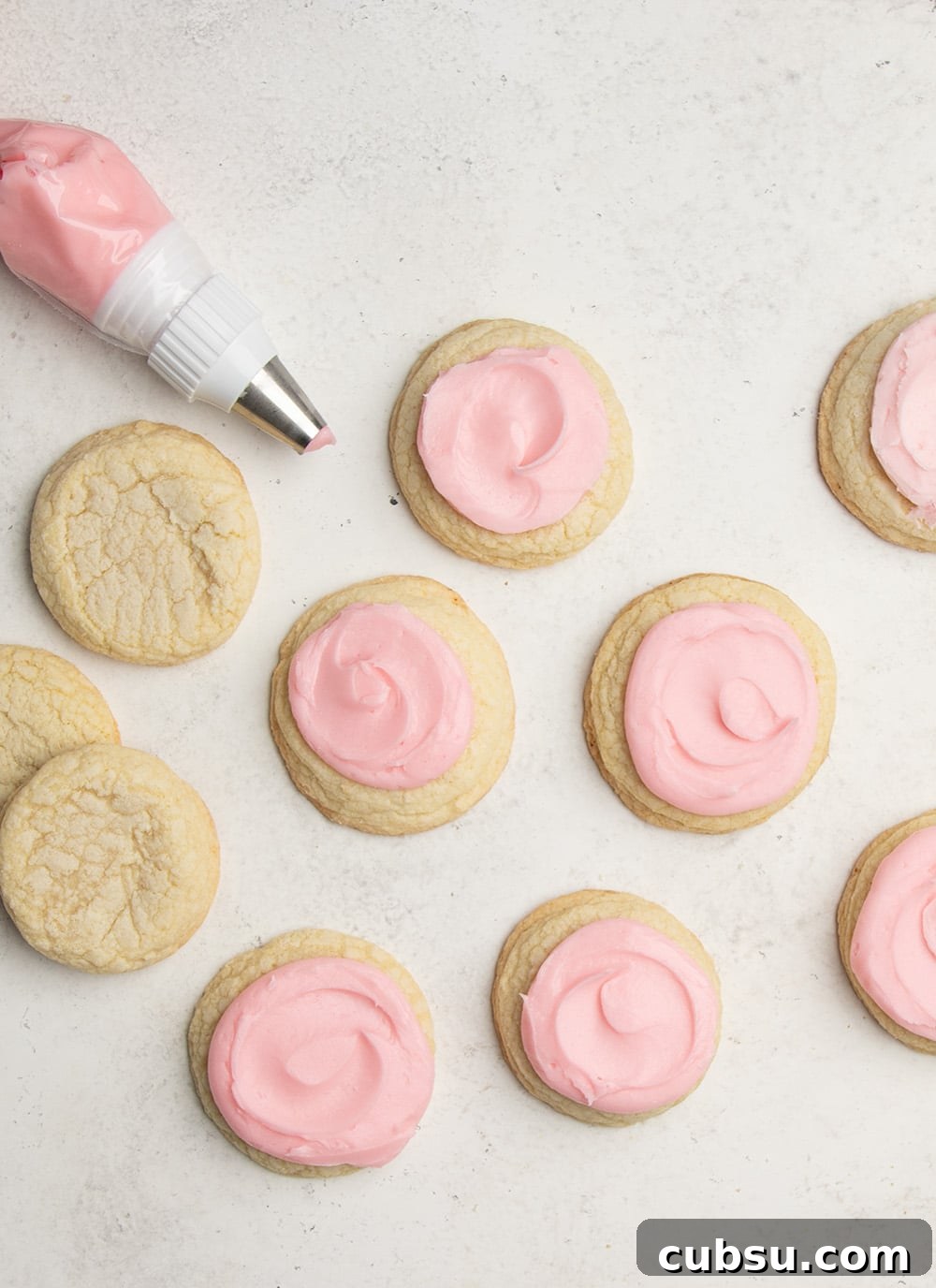 many light yellow sugar cookies on a white background, many with a swirl of pink frosting on top