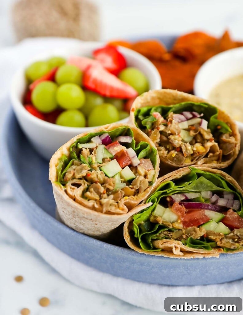 Three vegetable wraps in tortillas filled with lentils, tomato, cucumber, onion, and lettuce. There is a bowl of grapes and strawberries behind the wraps